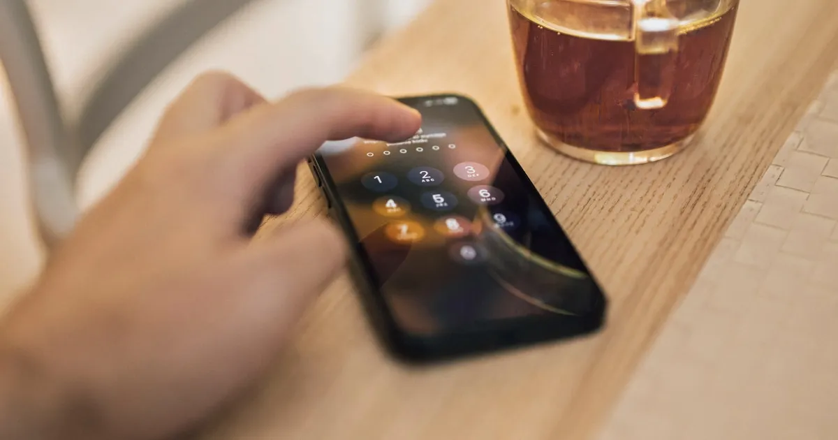 A hand enters the passcode on an iPhone lying on a wooden table, next to a glass.