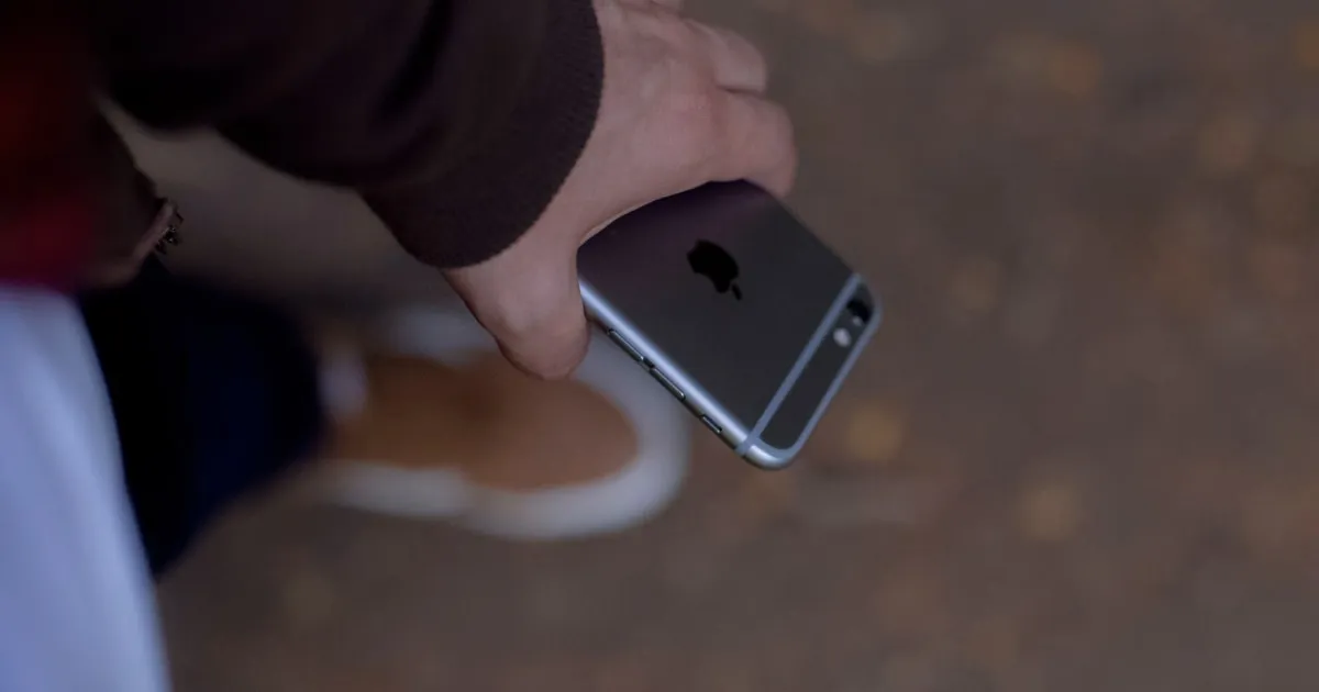 A hand grabs an iPhone held low to the ground, near a passerby’s shoes.