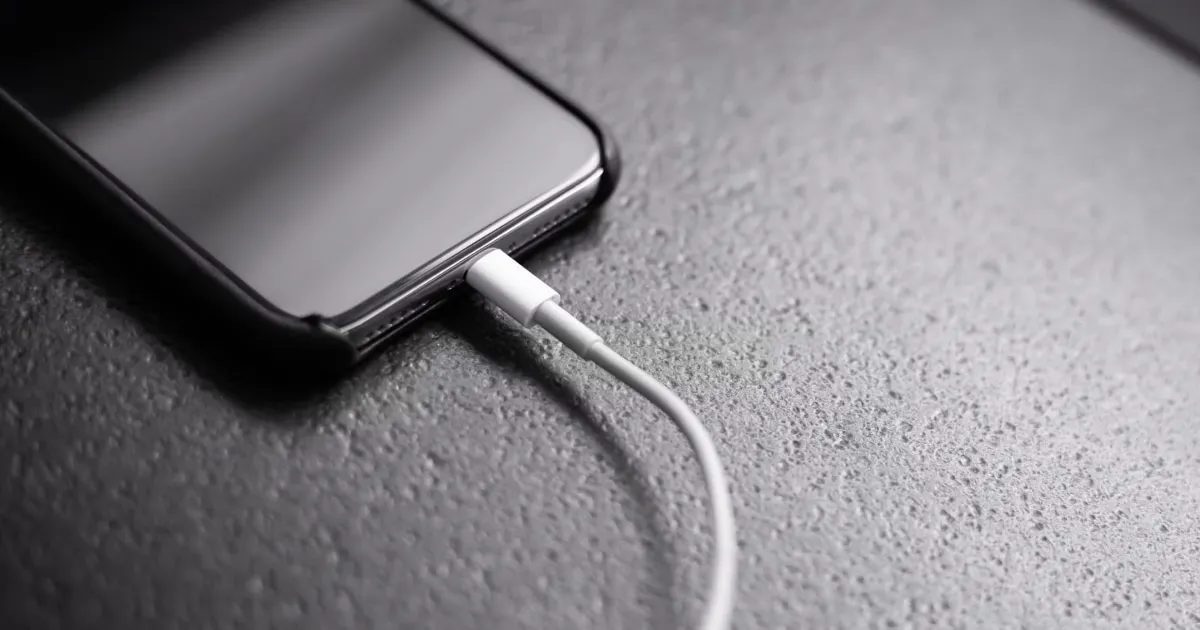 A black iPhone plugged into a white charging cable on a dark textured surface.