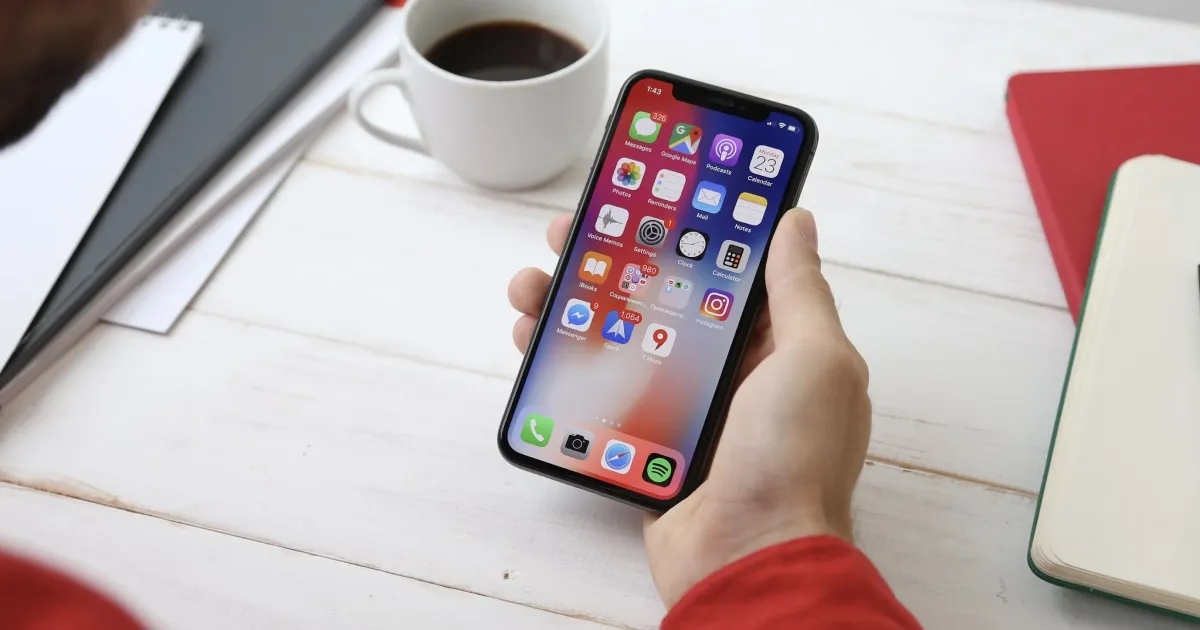 A hand holds an iPhone showing colorful apps, on a table with coffee and notebooks.