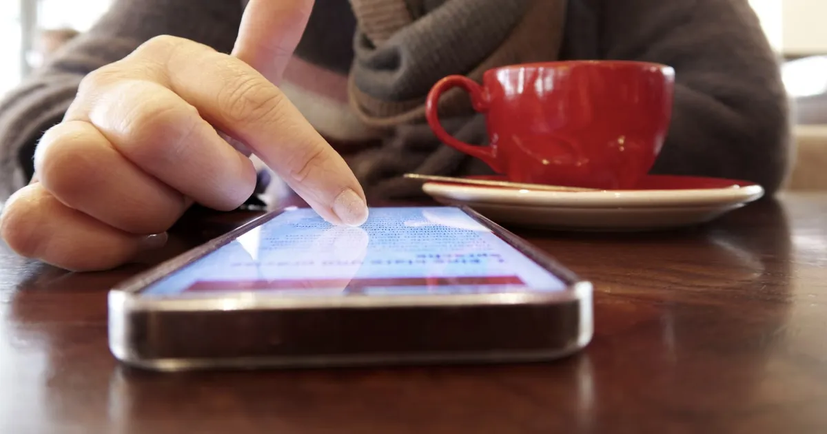 A finger touches the screen of an iPhone lying on a table, in front of a red cup.