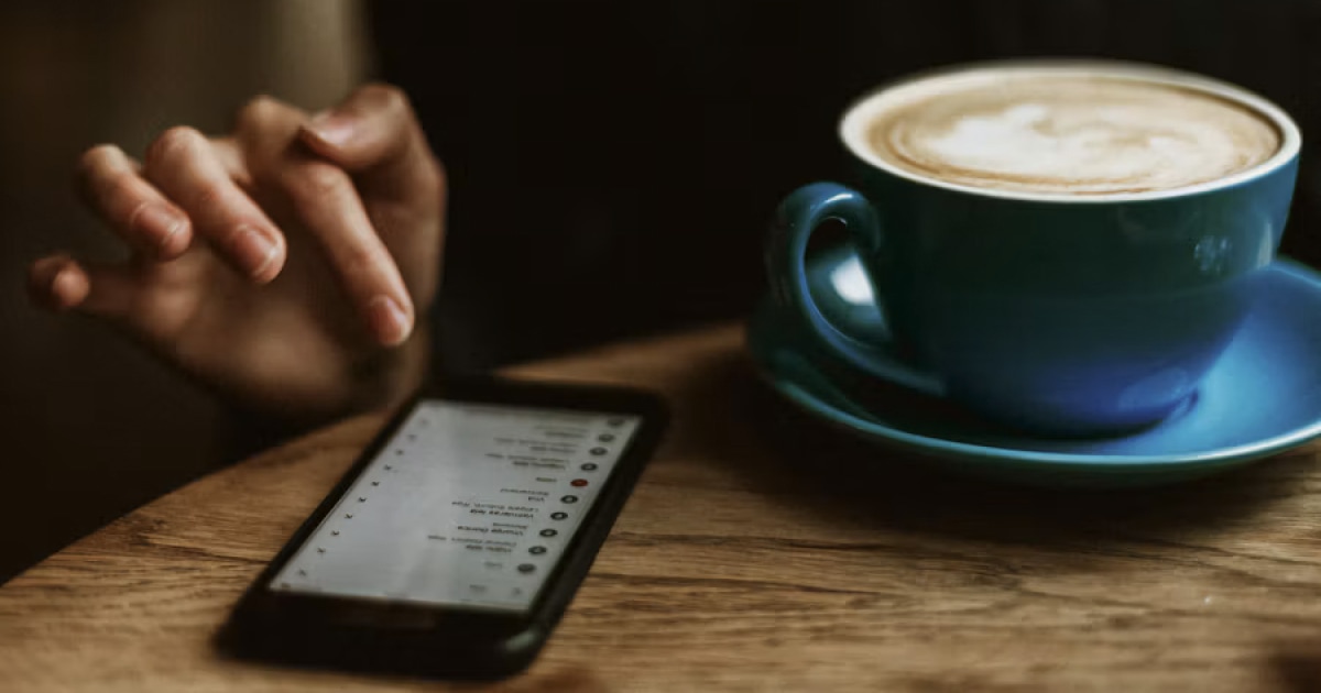 An iPhone showing a list of messages on a table, next to a cup of coffee.