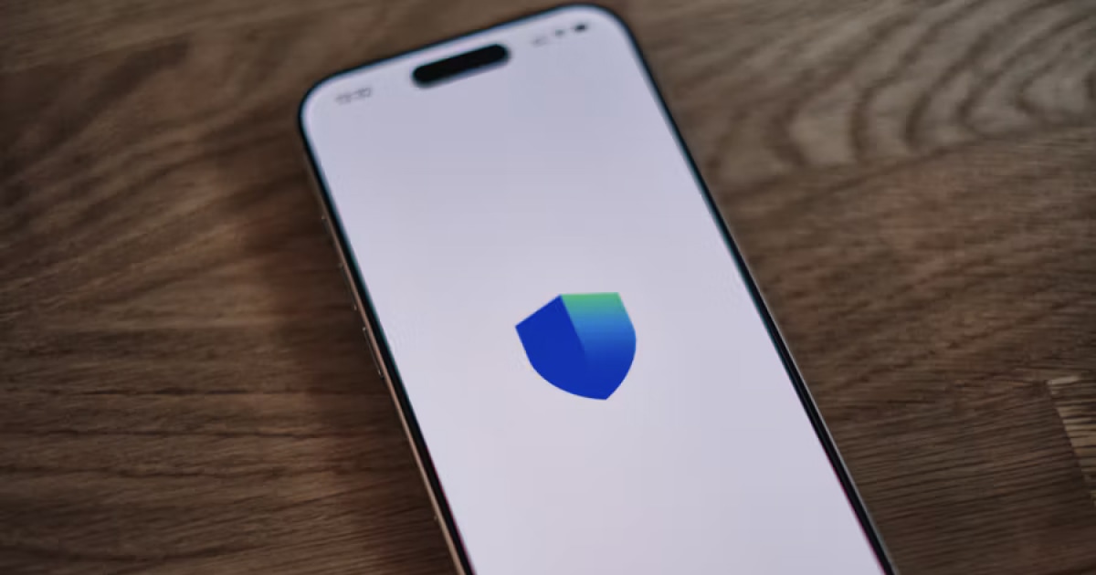 An iPhone on a wooden table displays a blue and green shield on its screen.