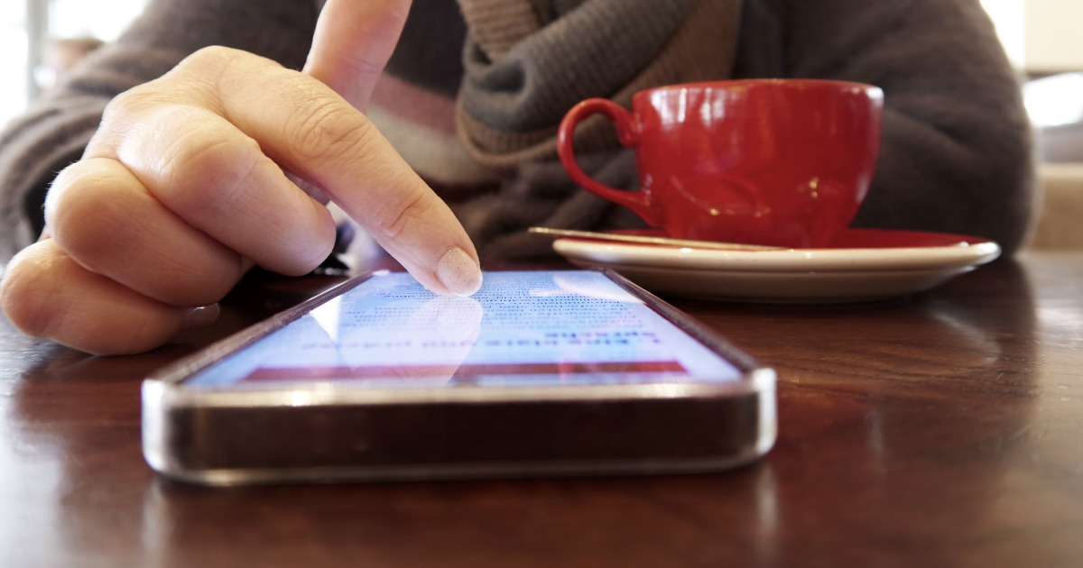 A finger touches the screen of an iPhone lying on a table, in front of a red cup.